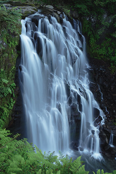 Before towering waterfall plunges over multiple layers of dark, weathered rocks