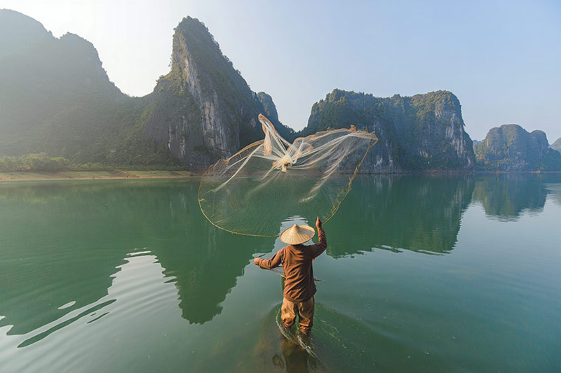 Before fisherman stands in ankle-deep water while casting a circular fishing net