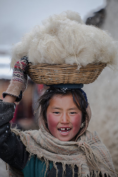 Before child in a Himalayan village balances a woven basket filled with white yak wool on her head